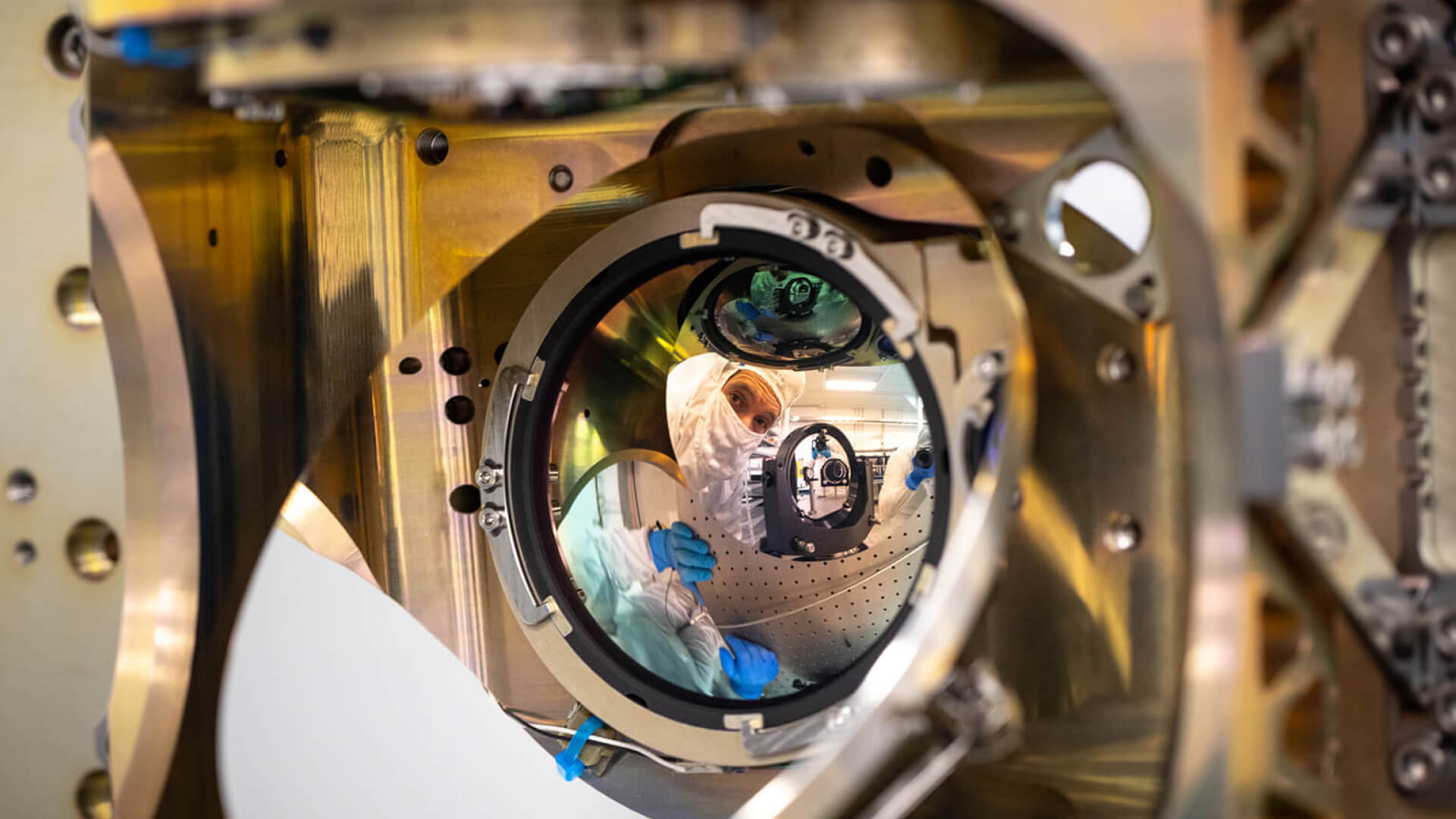 A cleanroom technician in a white suit and blue gloves peers through a large precision optical assembly during SDA Phase II