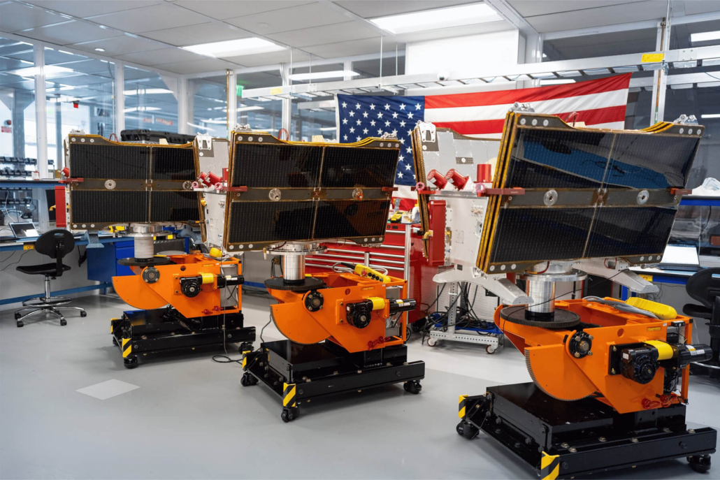 Three Vindler 2.0 satellites mounted on orange assembly stands in a cleanroom facility with an American flag in the