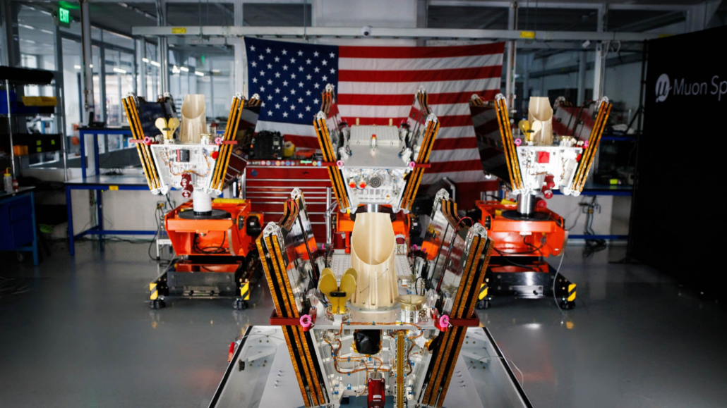 Multiple Muon Space satellites with deployed solar panel arms assembled in a factory facility with an American flag backdrop.