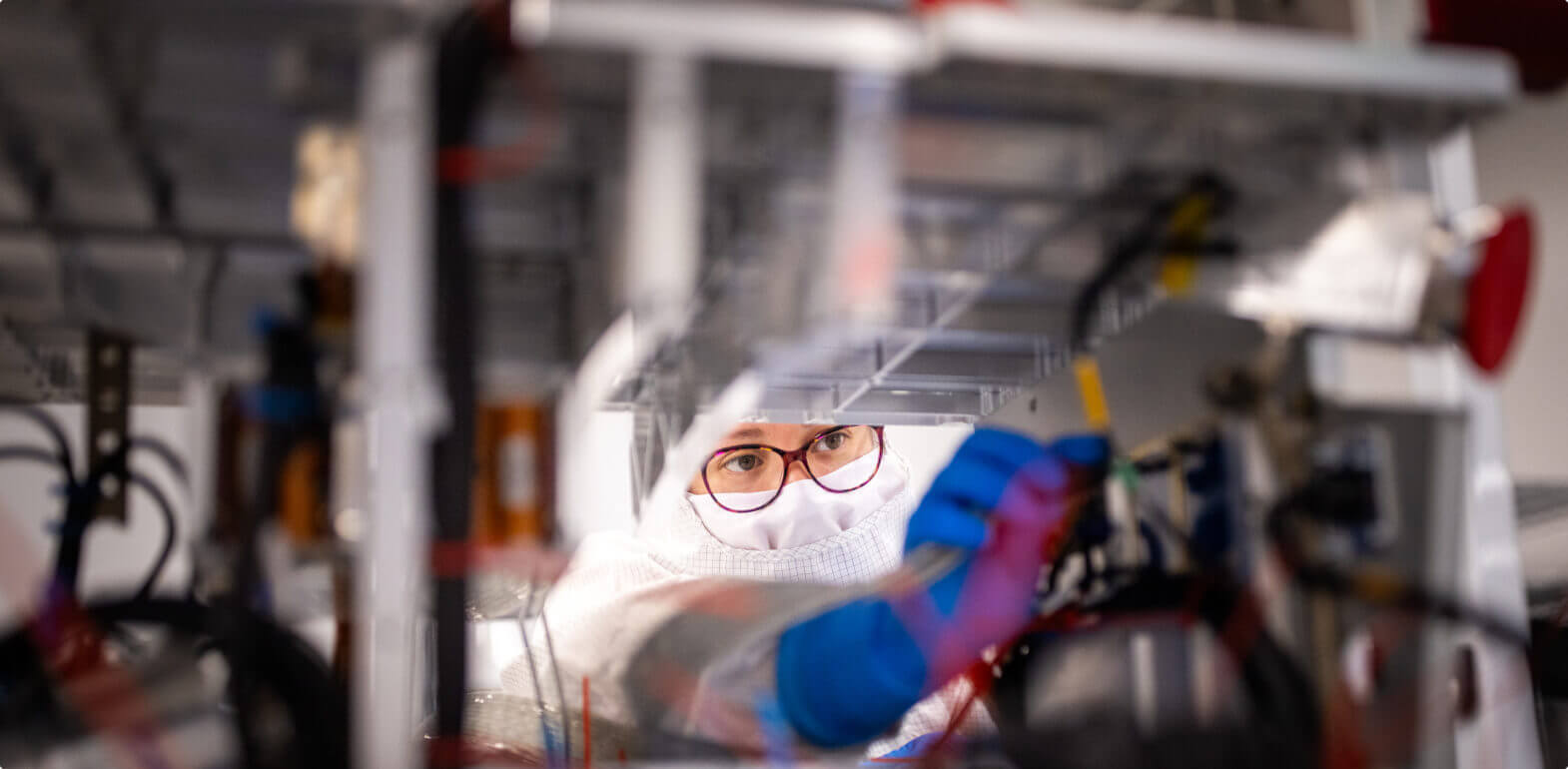 Engineer in cleanroom gear and blue gloves works on technical equipment surrounded by wires and components.