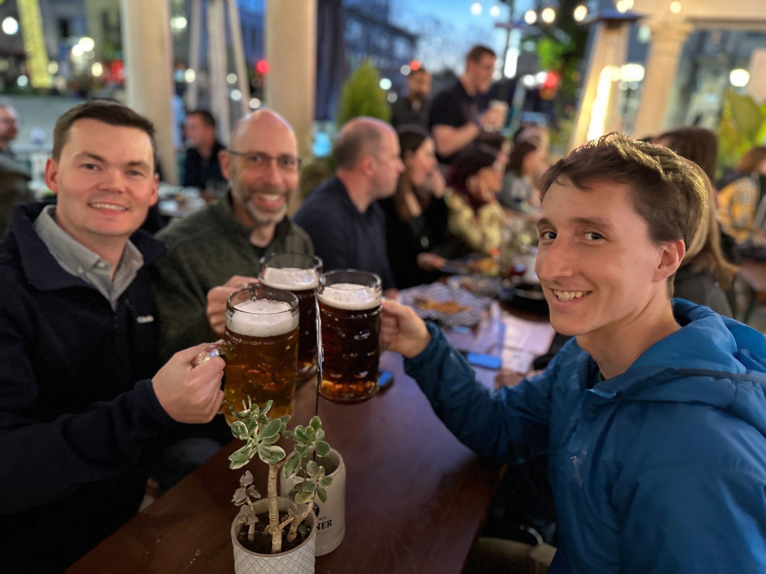 Muon Space team members smiling and clinking beer glasses together at an outdoor restaurant during an evening gathering.