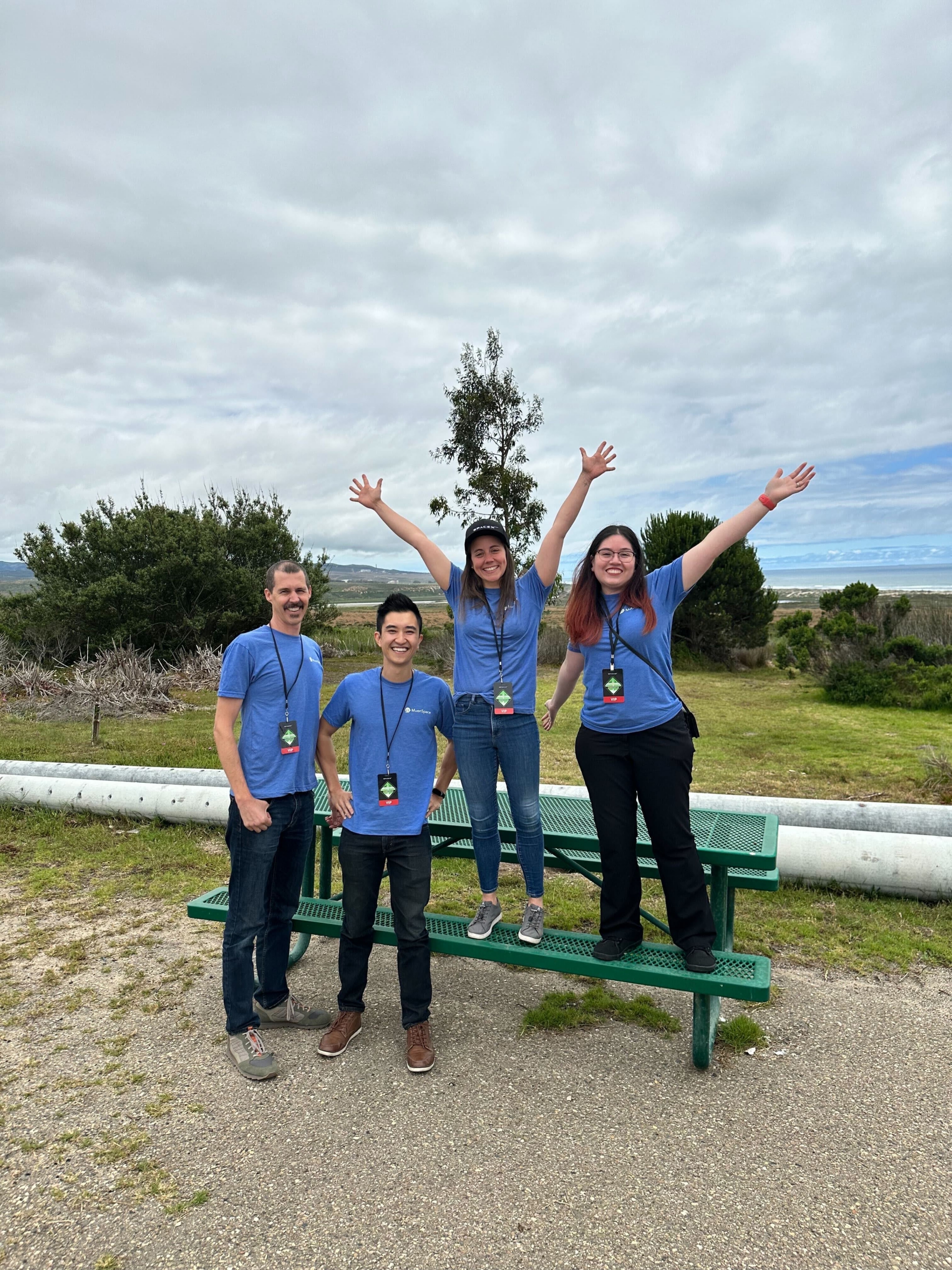 Four Muon Space team members in matching blue shirts pose cheerfully by a green picnic table at an outdoor launch site.