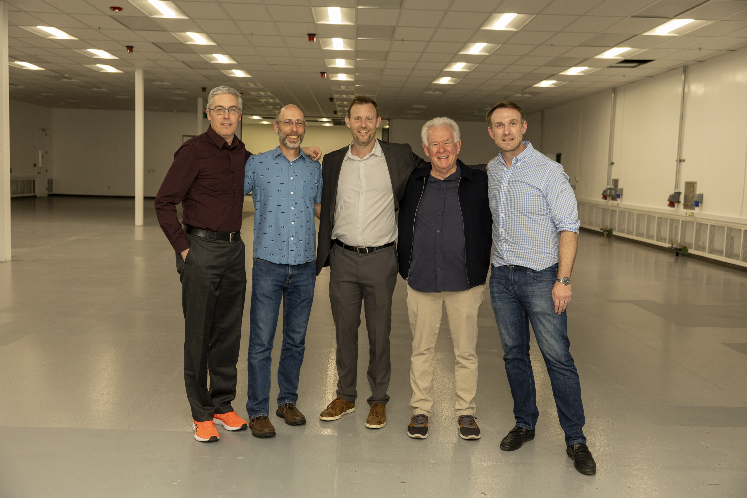Five smiling men pose together in a large, open industrial space during a Muon Space corporate party.
