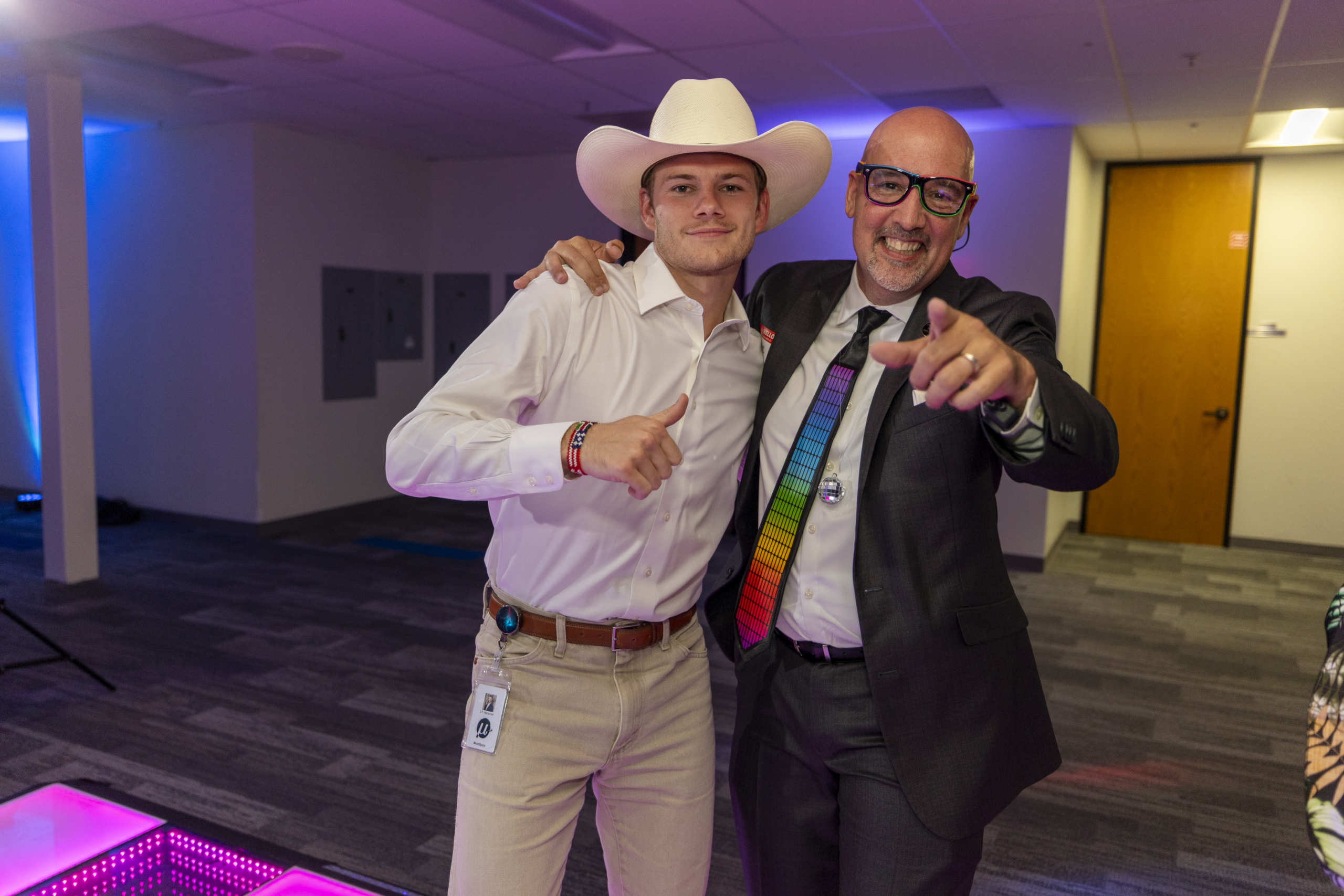 Two colleagues pose together at a corporate party, one wearing a cowboy hat and the other sporting a colorful LED tie and