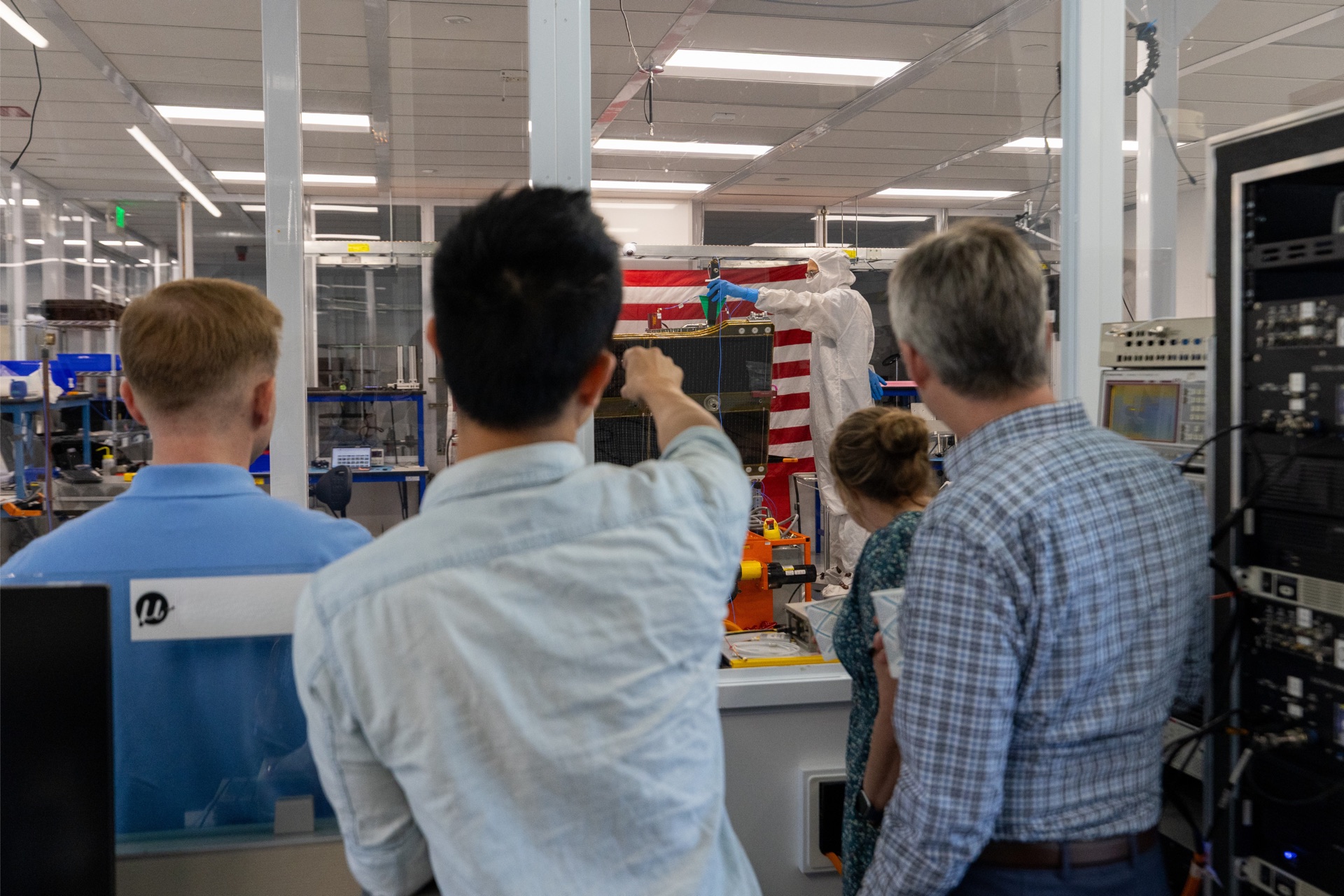 Visitors observe a technician in a cleanroom suit working on spacecraft hardware inside a clean room facility.