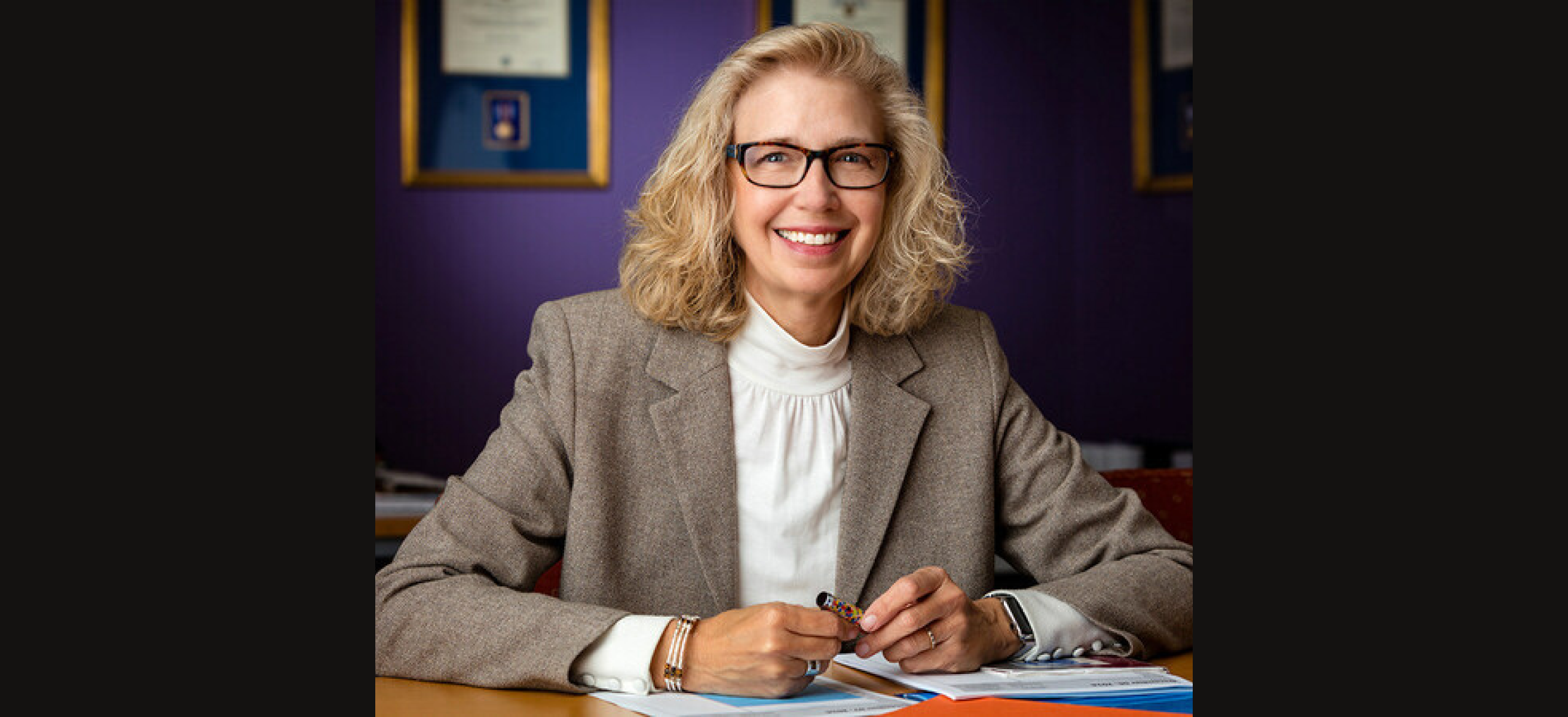Christine Fox smiling at a desk wearing glasses and a gray blazer, holding a pen with framed awards behind her.