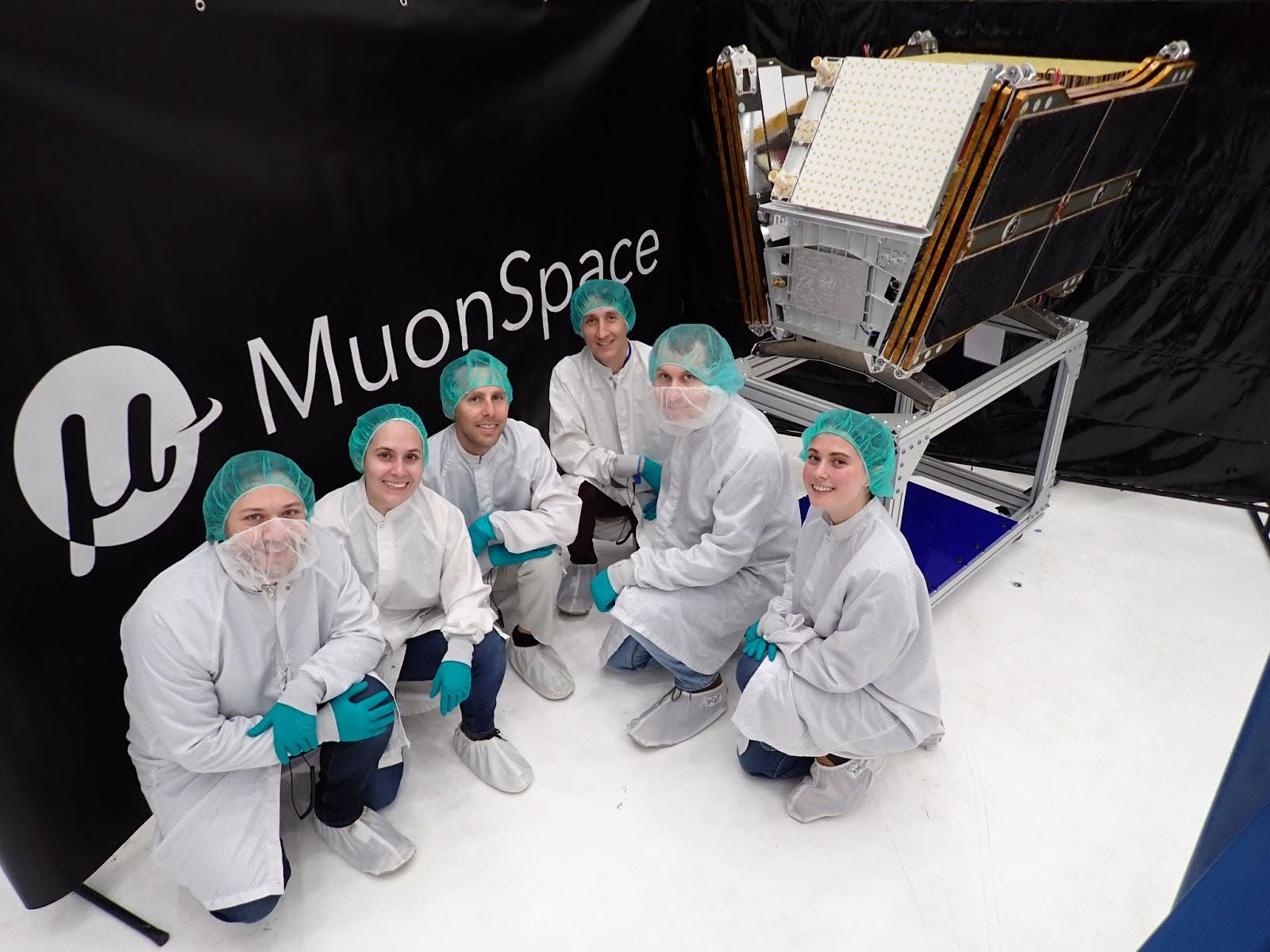 Muon Space team members in cleanroom suits pose beside the FireSat protoflight satellite in front of a branded banner.