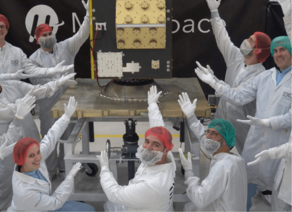 Muon Space team members in cleanroom suits and hairnets celebrate with raised hands around a satellite in a cleanroom