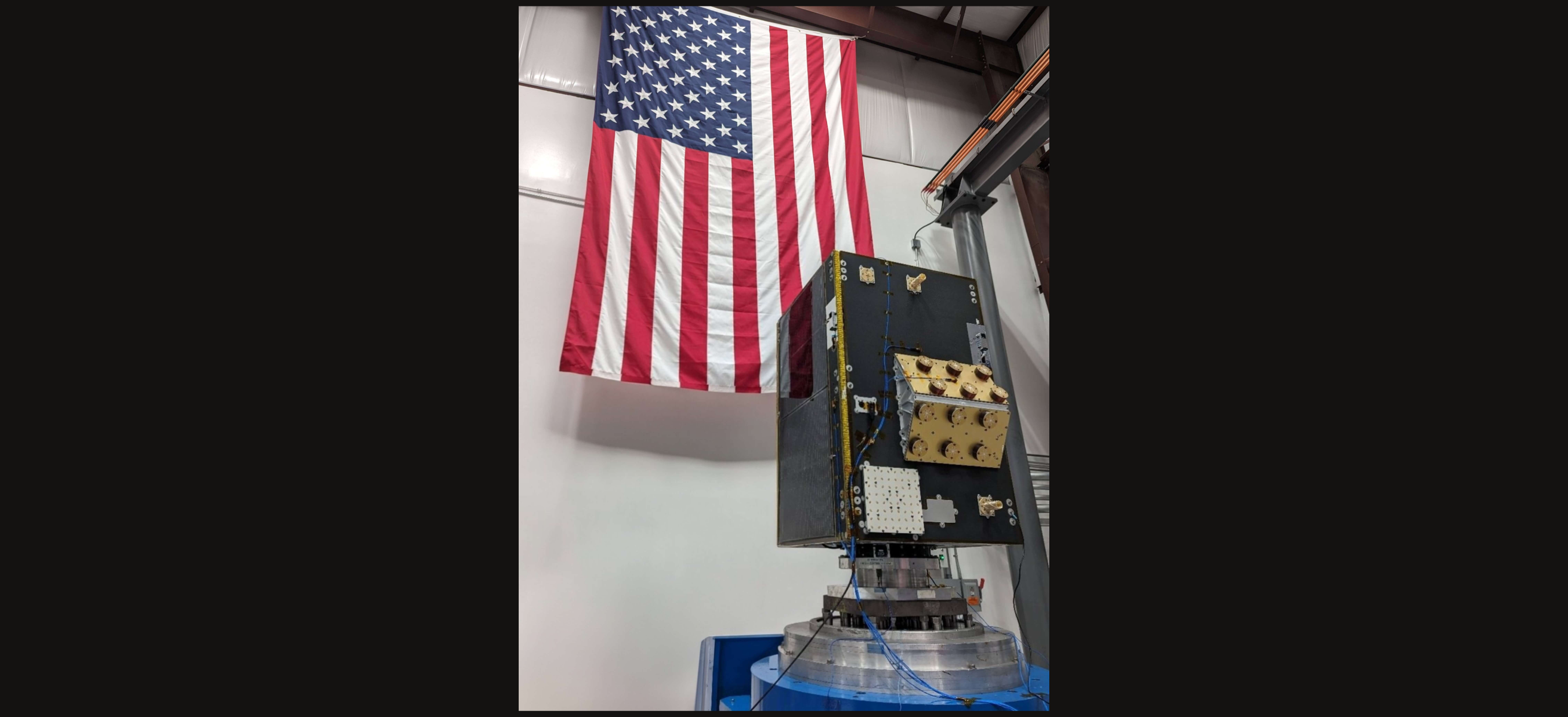 A commercial Earth observation satellite mounted on a test stand in a clean room with a large American flag hanging behind