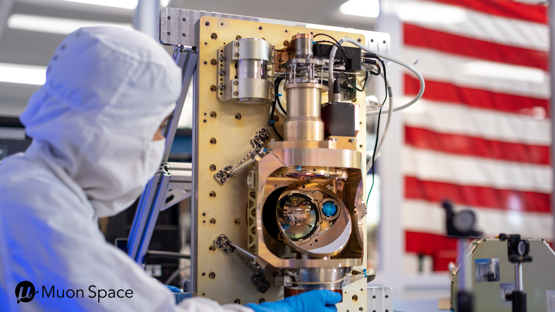 A cleanroom technician inspects a Muon Space satellite sensor payload with precision optics, with an American flag in the