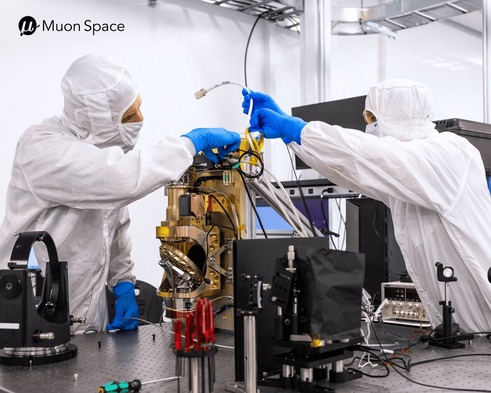 Two Muon Space engineers in cleanroom suits and blue gloves assemble satellite hardware on a lab workbench.
