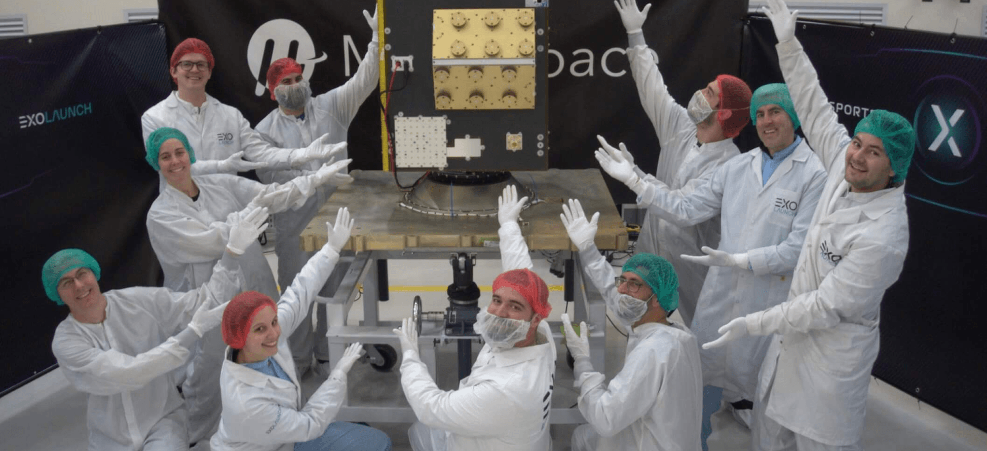 A team of engineers in cleanroom suits and hairnets celebrate with raised hands around the MuSat2 weather satellite.