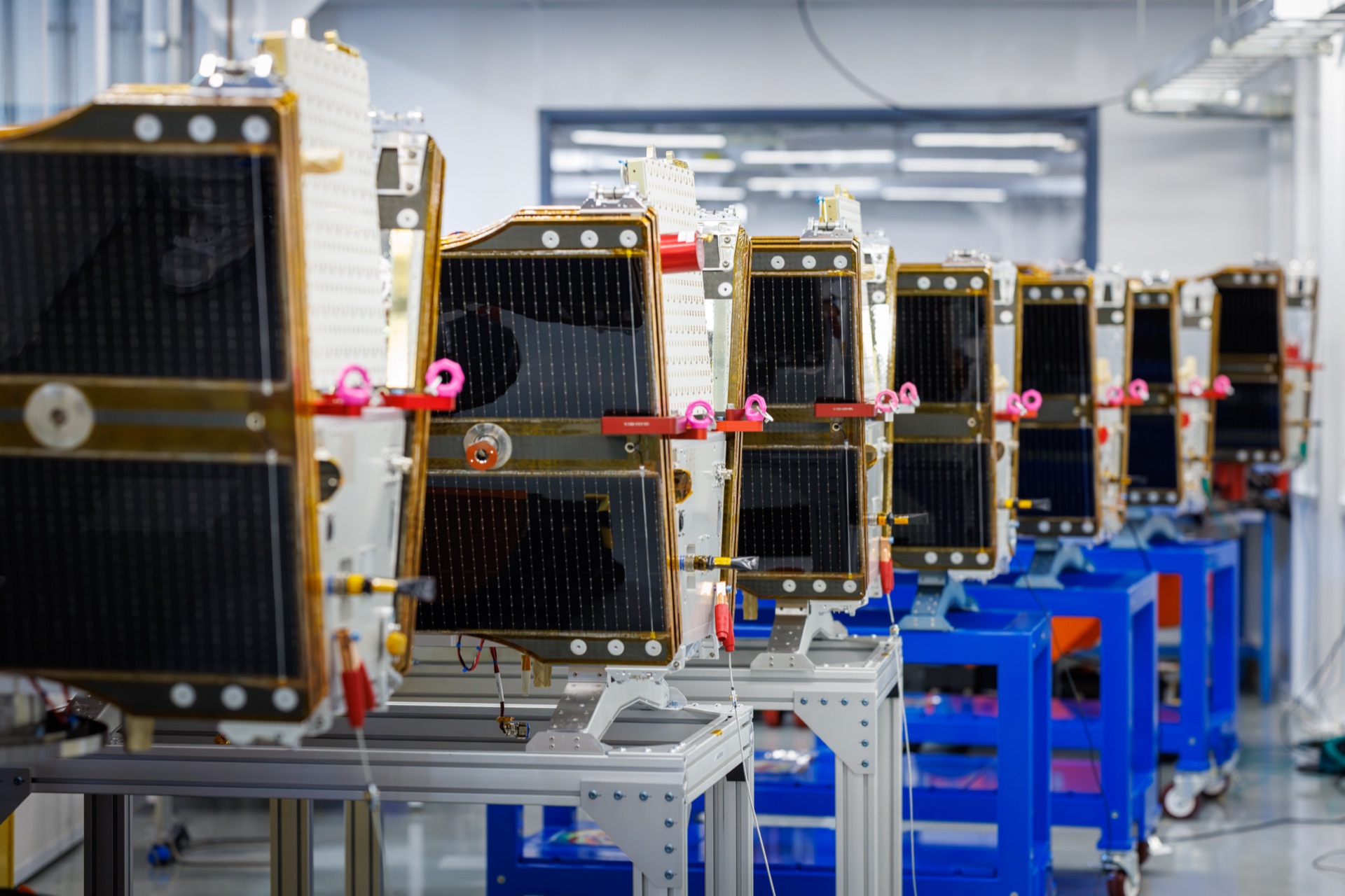 Multiple small satellites with gold-trimmed solar panels are lined up on assembly stands in a cleanroom facility.