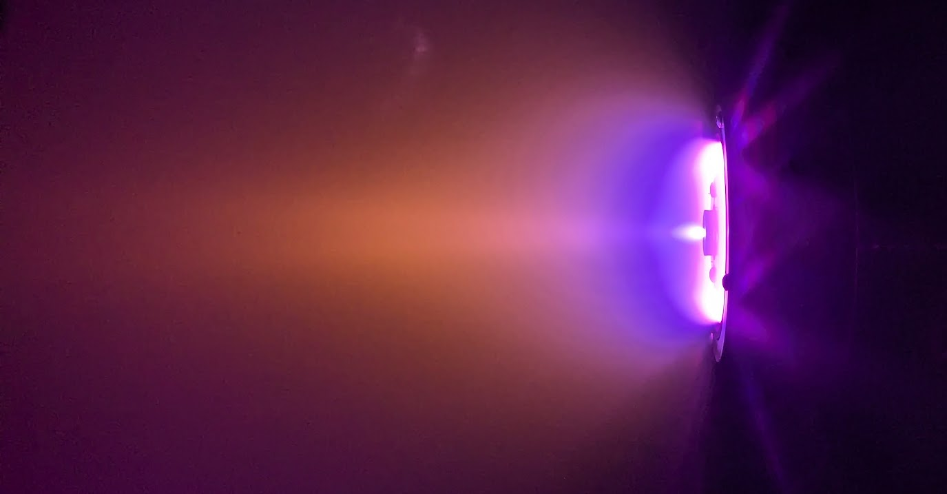 A plasma thruster emits a glowing purple and orange exhaust plume in a dark vacuum chamber during a test firing.